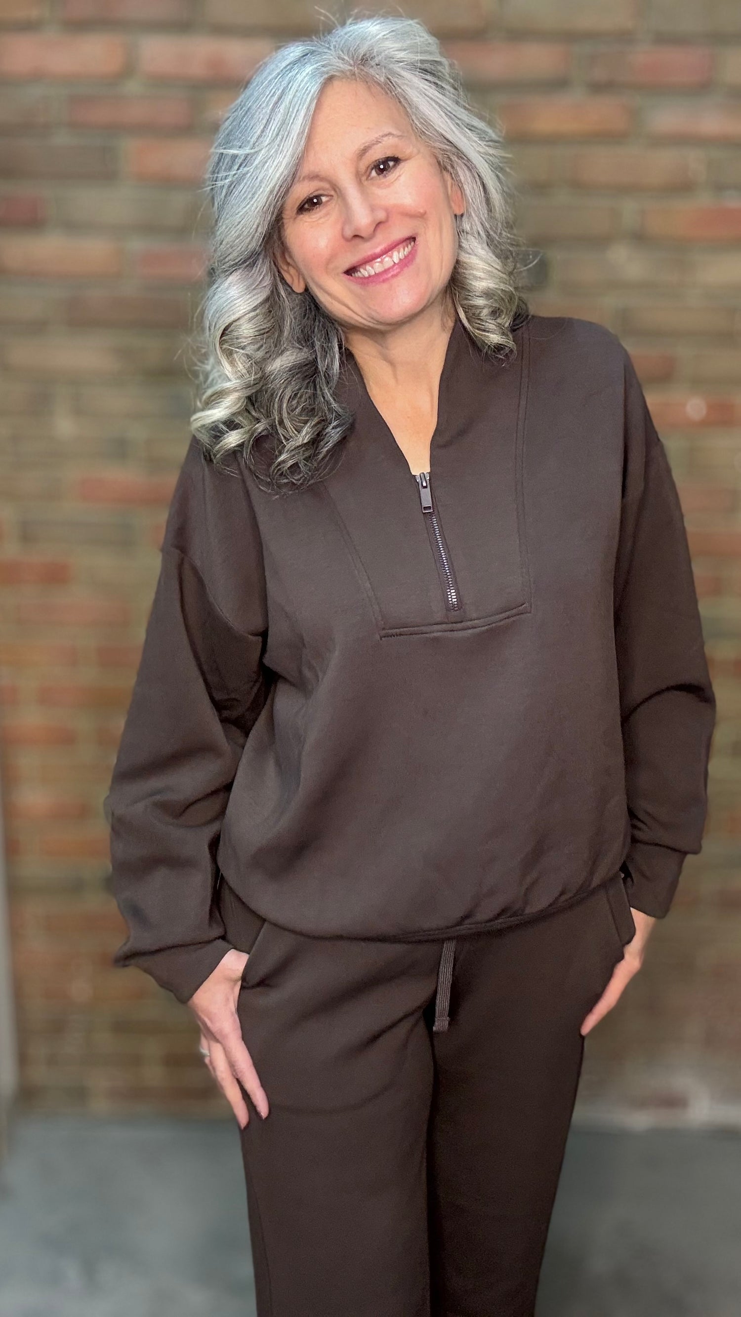 Woman wearing a brown pullover with a zipper in front of a brick wall.