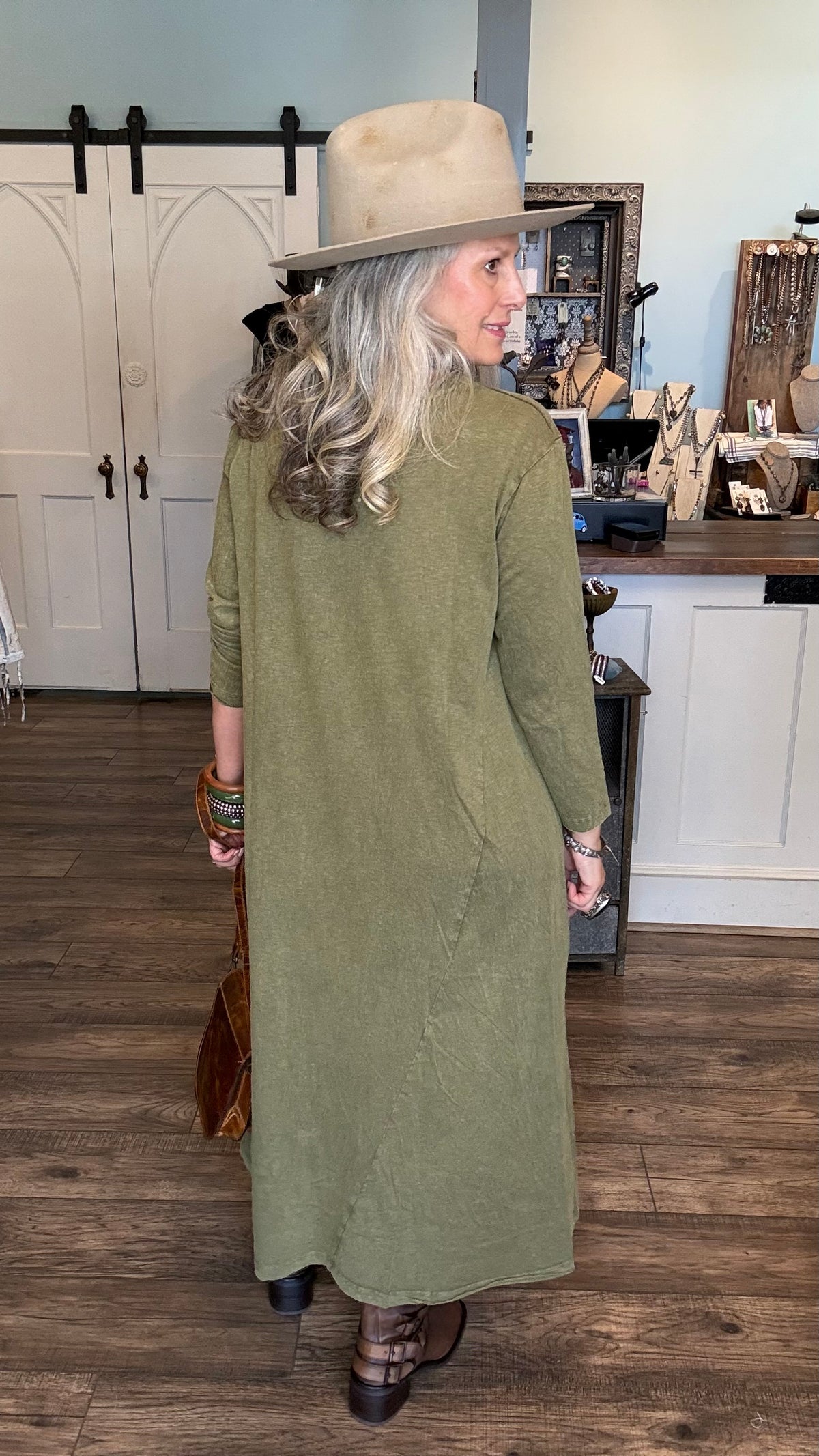 A woman wearing a long-sleeved olive green maxi dress, a beige wide-brim hat, and brown shoes, standing in a rustic shop with wood floors and white barn-style doors.