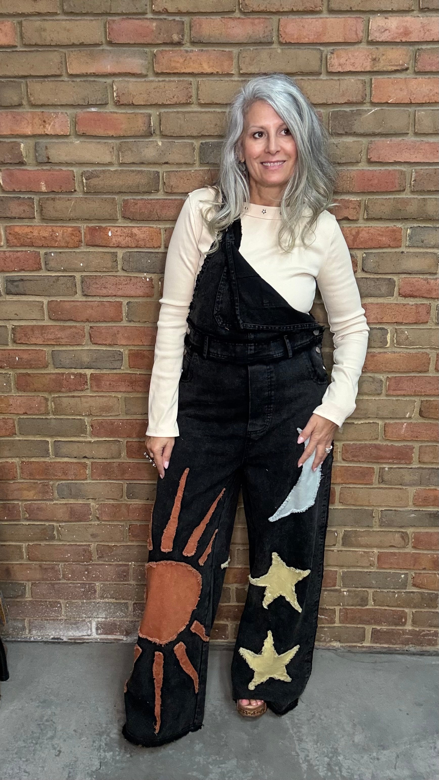 A woman with gray hair stands against a brick wall wearing black denim overalls with orange sun and gold star patches, layered over a cream asymmetrical top.