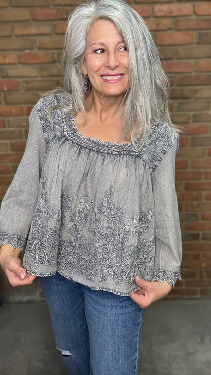 Woman with gray hair wearing the Vintage Washed Floral Lace Top a  light gray blouse and blue jeans standing against a brick wall.