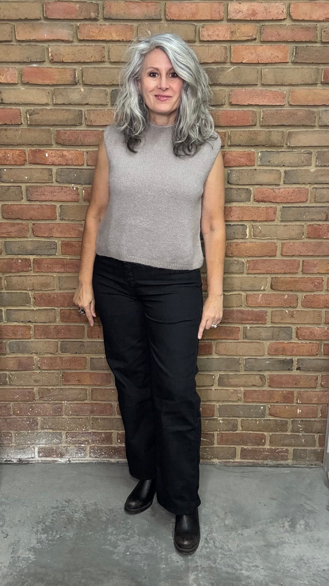 Person wearing a gray sleeveless top and black pants standing against a brick wall.