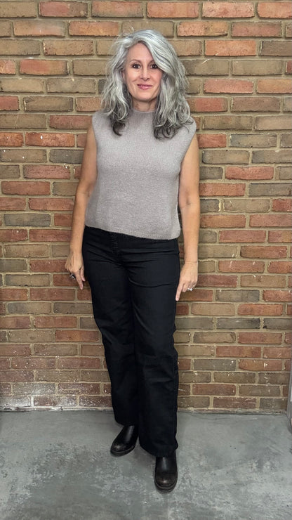 Person wearing a gray sleeveless top and black pants standing against a brick wall.