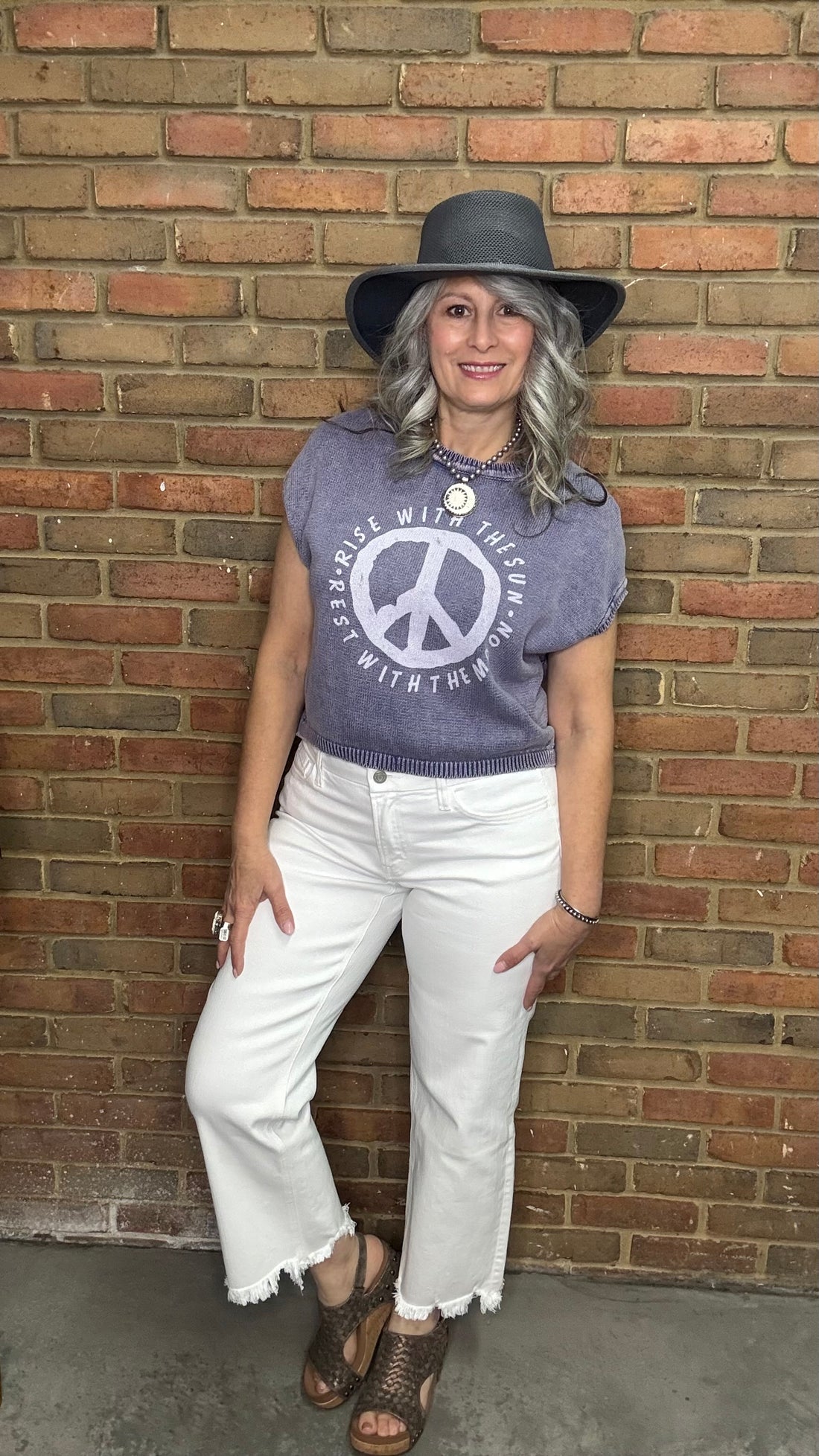 Woman wearing a Purple shirt with Peace Sign and Mid Rise Uneven Hem Crop Jeans against a brick wall.