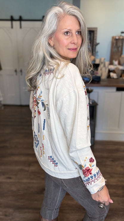 A woman with long gray hair is shown from the back wearing a cream-colored embroidered jacket with colorful floral and geometric patterns, paired with gray pants in an indoor home setting.