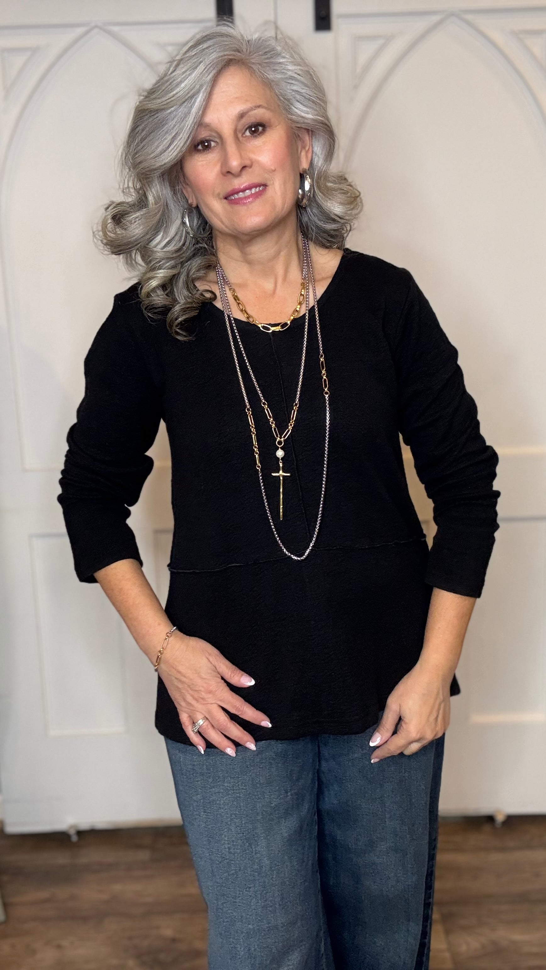 Woman wearing The Black Wanda Round Neck Top layered with the Silver Snake and Gold Paperclip Chain Necklace, the Silver Snake and Gold Paperclip Chain Choker Necklace and the Gold Cross w/Pearl Necklace  paired with  Judy Blue Palazzo jeans indoors against a white paneled wall.