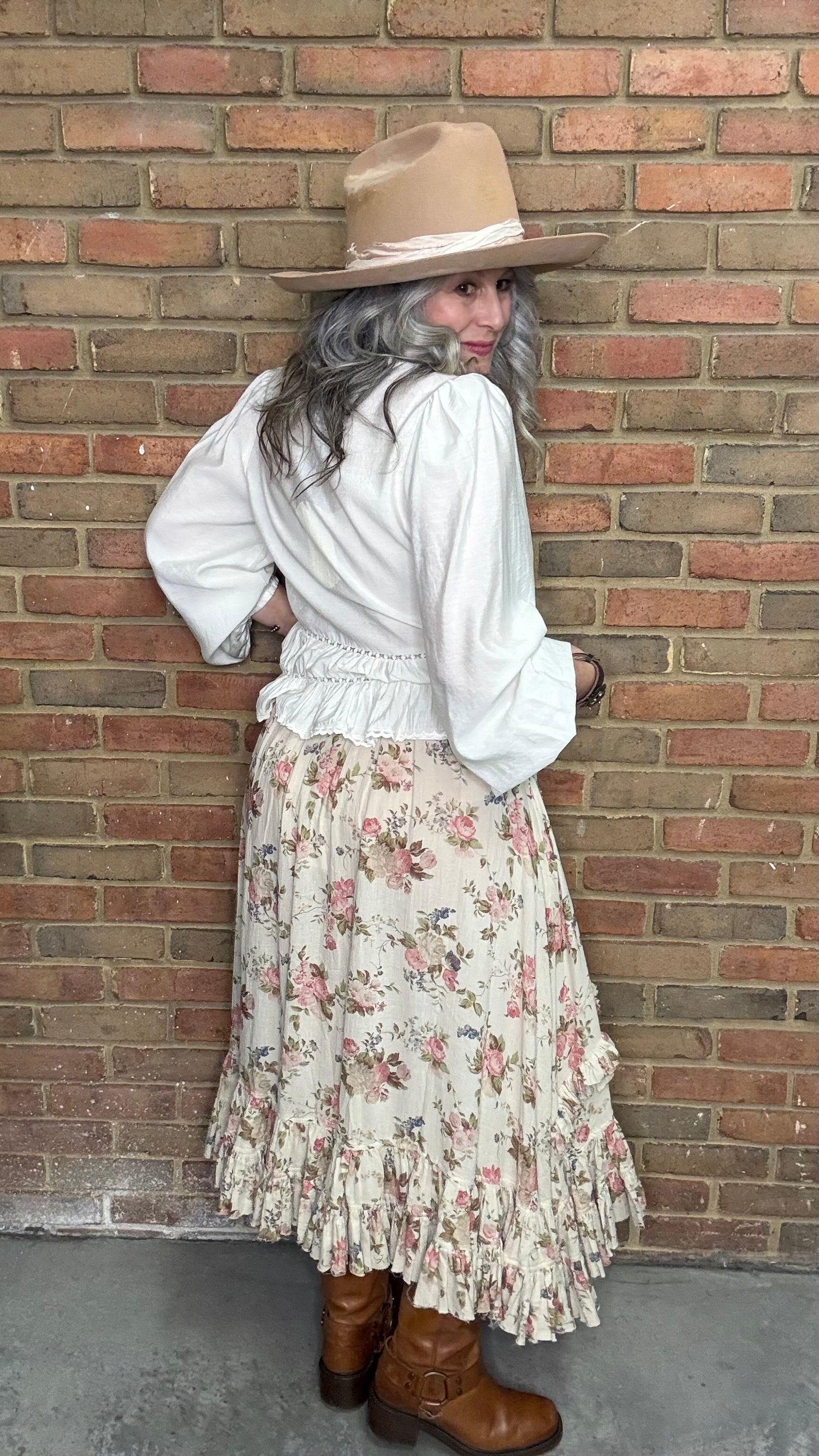 Woman in a White Pin-Tuck Lace Embellished shirt, blooming romance skirt, and Kate Harness Frye boots standing against a brick wall.