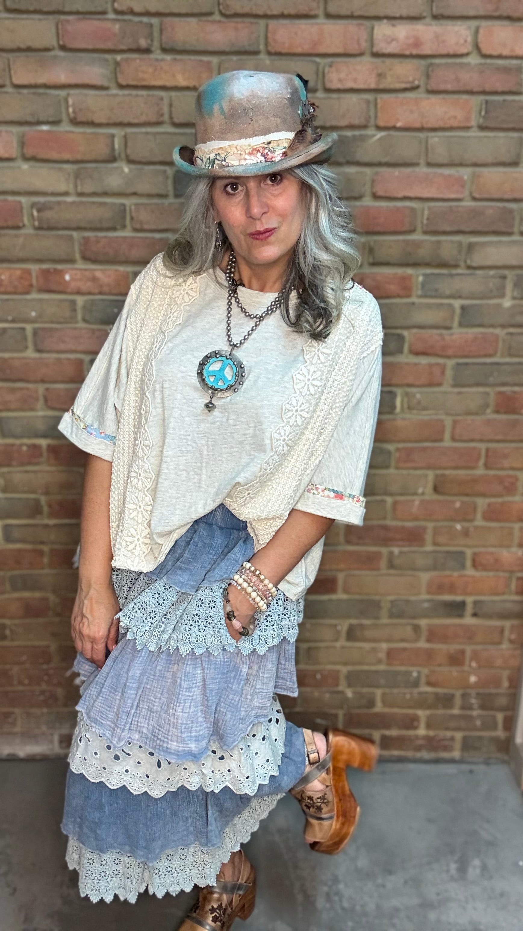 A woman wearing a bohemian-style outfit consisting of a cream lace top, a blue tiered pant, a brown hat, and layered necklaces, standing against a brick wall.