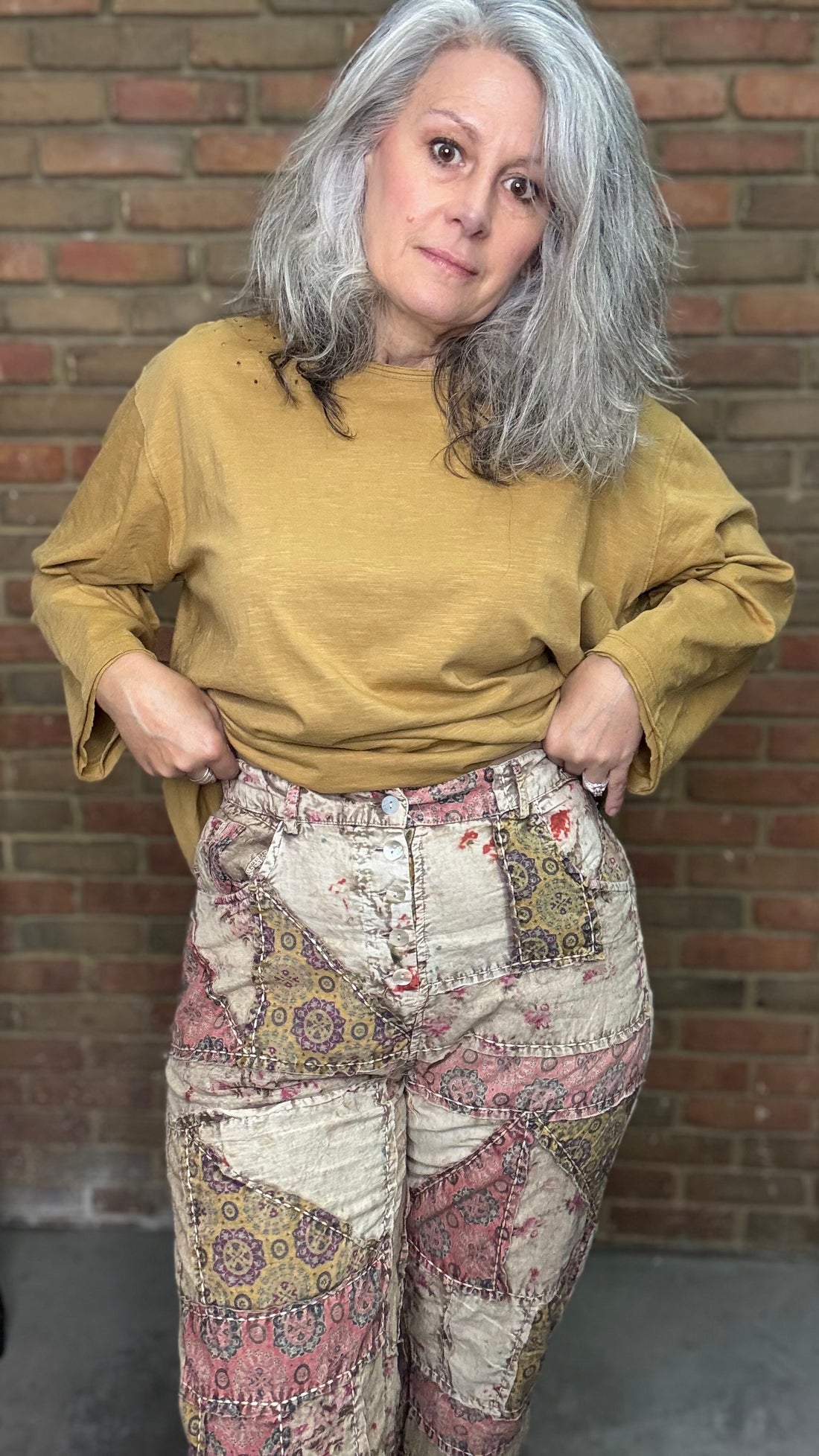 Woman wearing a mustard yellow sweater and patterned pants standing against a brick wall.
