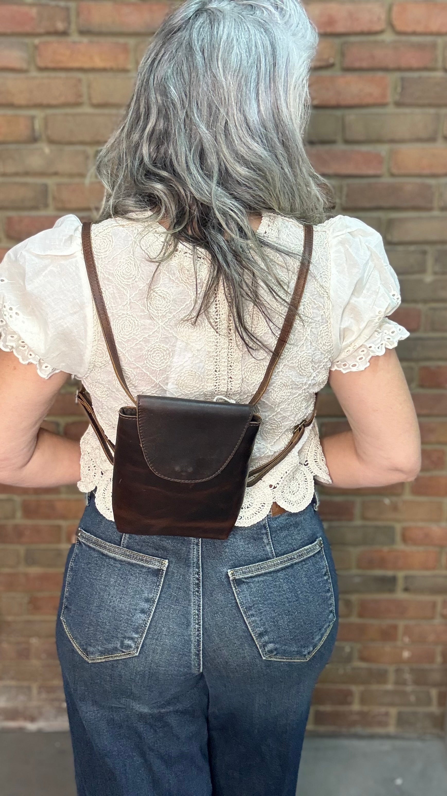 Woman holding a Backpack style leather Handbag with a brick wall in background.