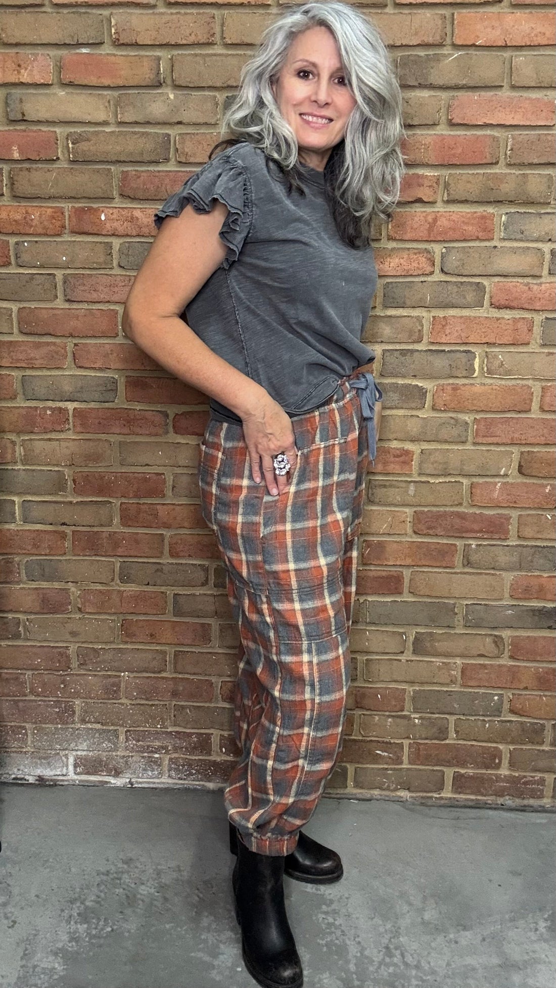 Woman in plaid pants and gray top leaning against a brick wall