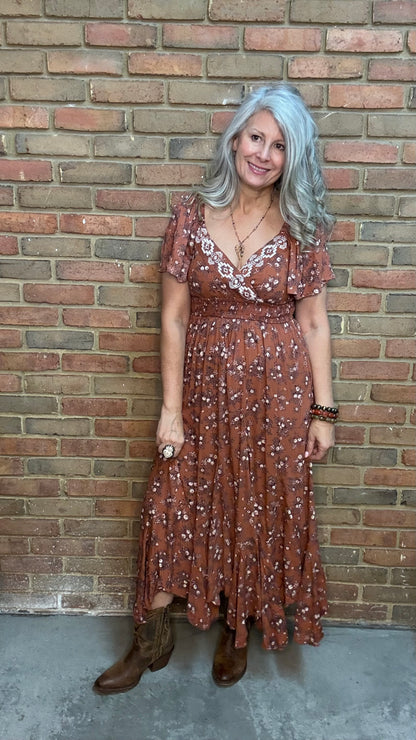 A woman with gray hair stands against a brick wall wearing a rust-brown bohemian-style midi dress with white floral prints, the "Short Sleeve Embroidered Midi Dress" and brown cowboy boots.