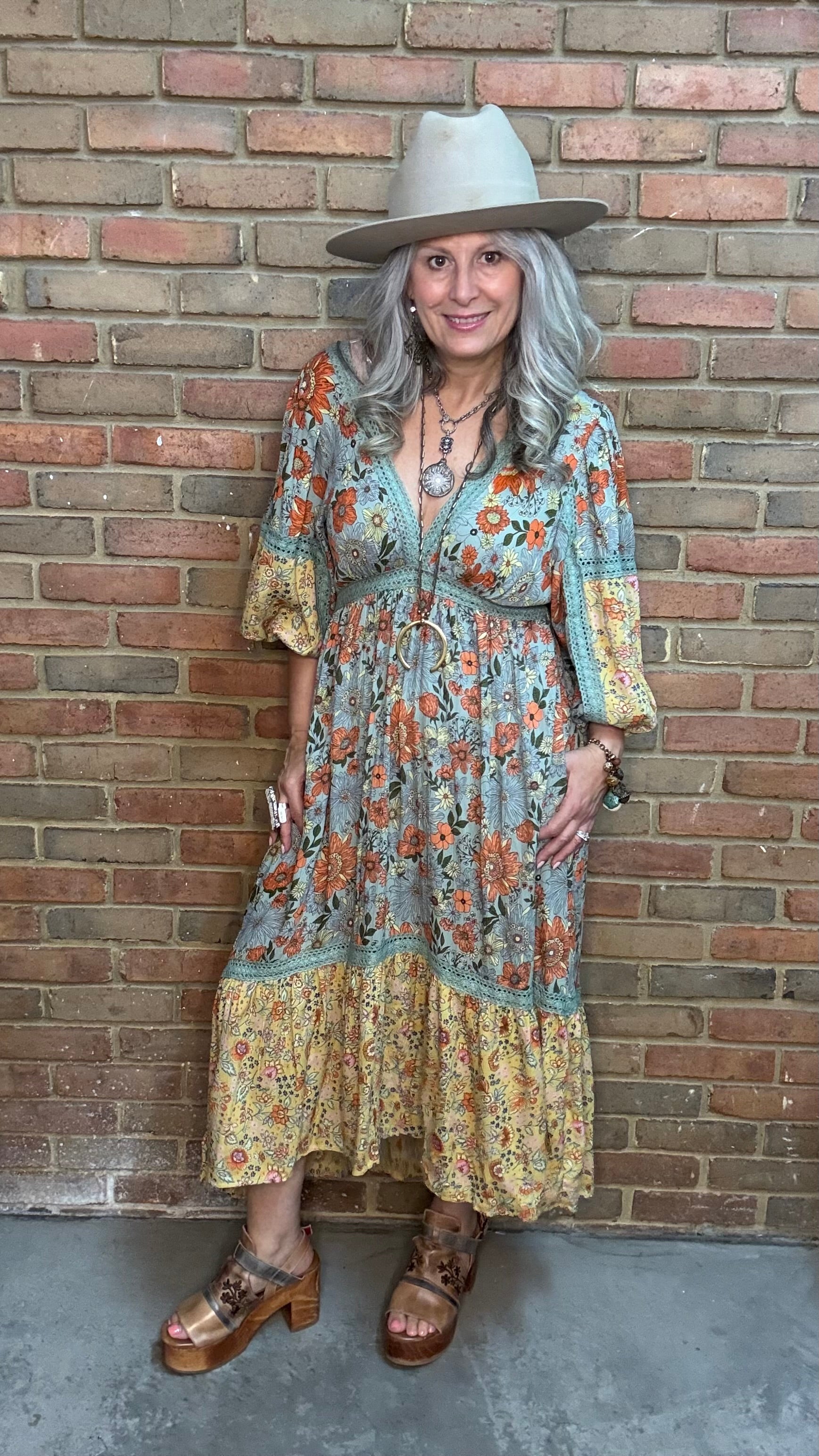 A woman stands against a brick wall wearing a bohemian-style midi dress, the  Long Sleeve V-Neck Hi-Lo Dress, with a blue-gray base and orange floral print, a wide-brimmed beige hat, and brown platform sandals.