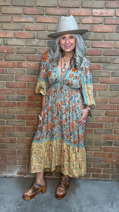 A woman stands against a brick wall wearing a bohemian-style midi dress, the  Long Sleeve V-Neck Hi-Lo Dress, with a blue-gray base and orange floral print, a wide-brimmed beige hat, and brown platform sandals.