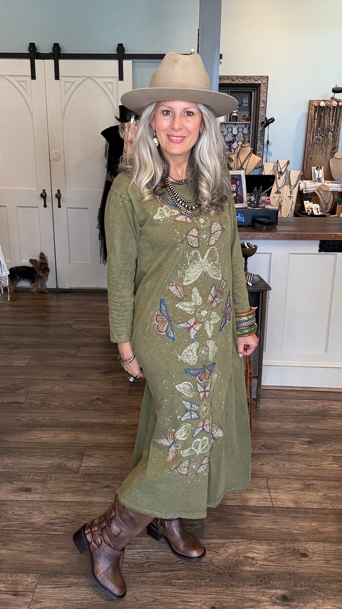 A woman wearing a long-sleeve olive green maxi dress with colorful butterfly embroidery, a beige wide-brim hat, and brown leather boots, standing in a rustic indoor boutique setting with wood floors and white barn-style doors in the background.