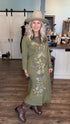 A woman wearing a long-sleeve olive green maxi dress with colorful butterfly embroidery, a beige wide-brim hat, and brown leather boots, standing in a rustic indoor boutique setting with wood floors and white barn-style doors in the background.