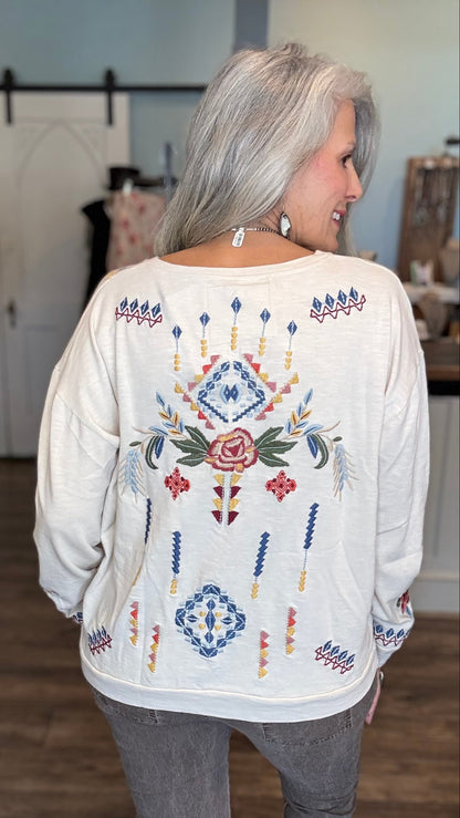 A woman with silver hair is shown from behind wearing a cream-colored long-sleeve top with a colorful embroidered geometric and floral pattern on the back, paired with gray pants and brown boots in an indoor boutique setting.