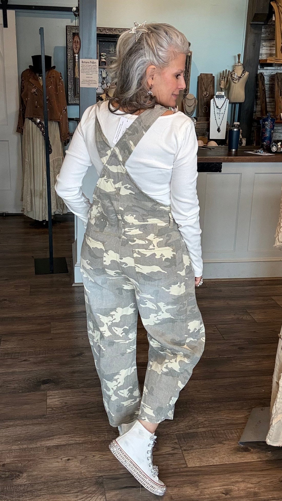 A woman with silver hair tied back is wearing gray and beige camouflage overall over a white long-sleeve shirt, standing in a boutique with wood floors and clothing racks.