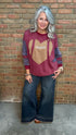 A woman with silver hair stands against a brick wall wearing a long-sleeve crewneck sweatshirt with a football graphic and dark wide-leg jeans.
