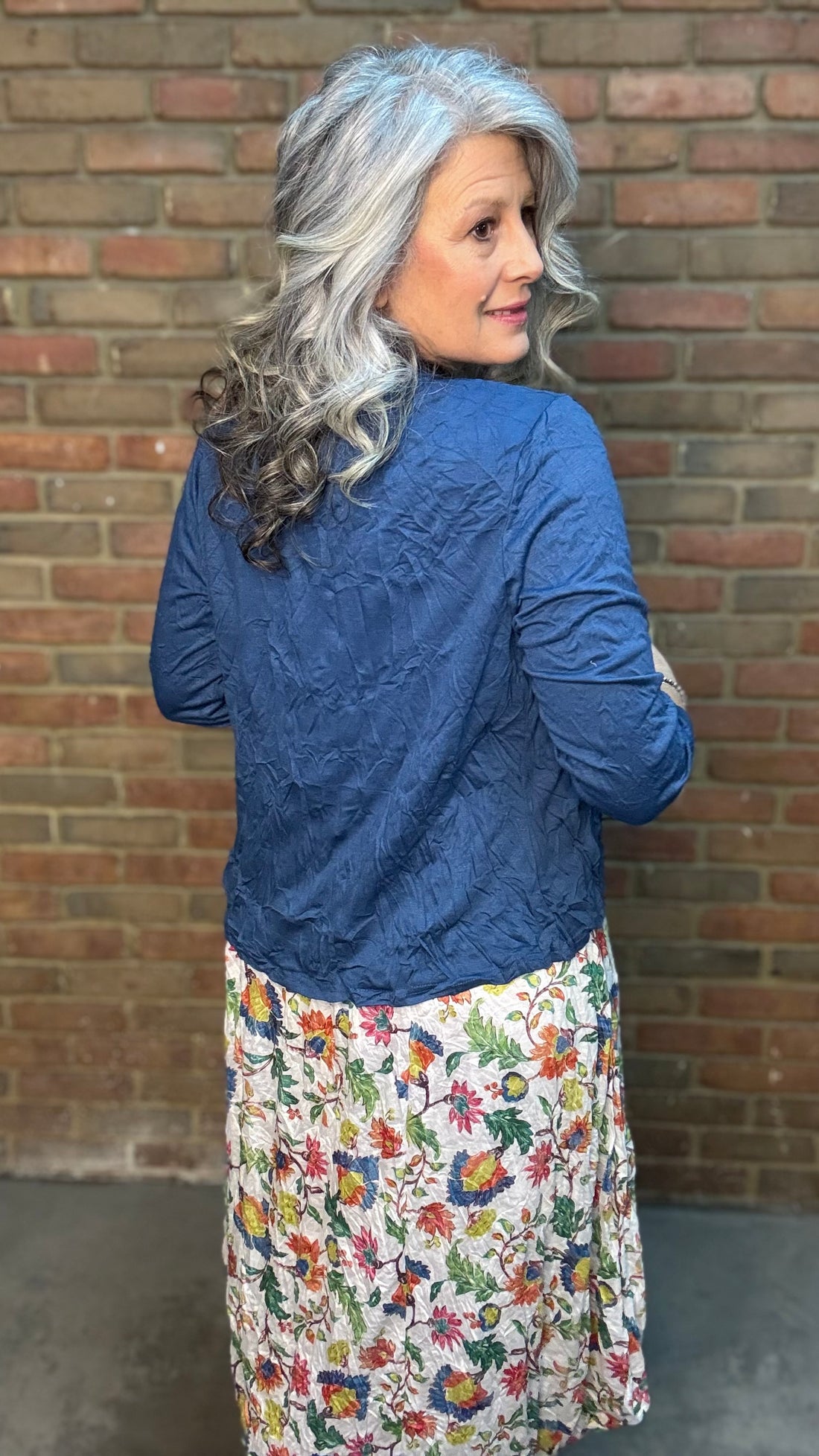 Woman with gray hair wearing Indigo Crinkle Cardigan over a floral dress against a brick wall.
