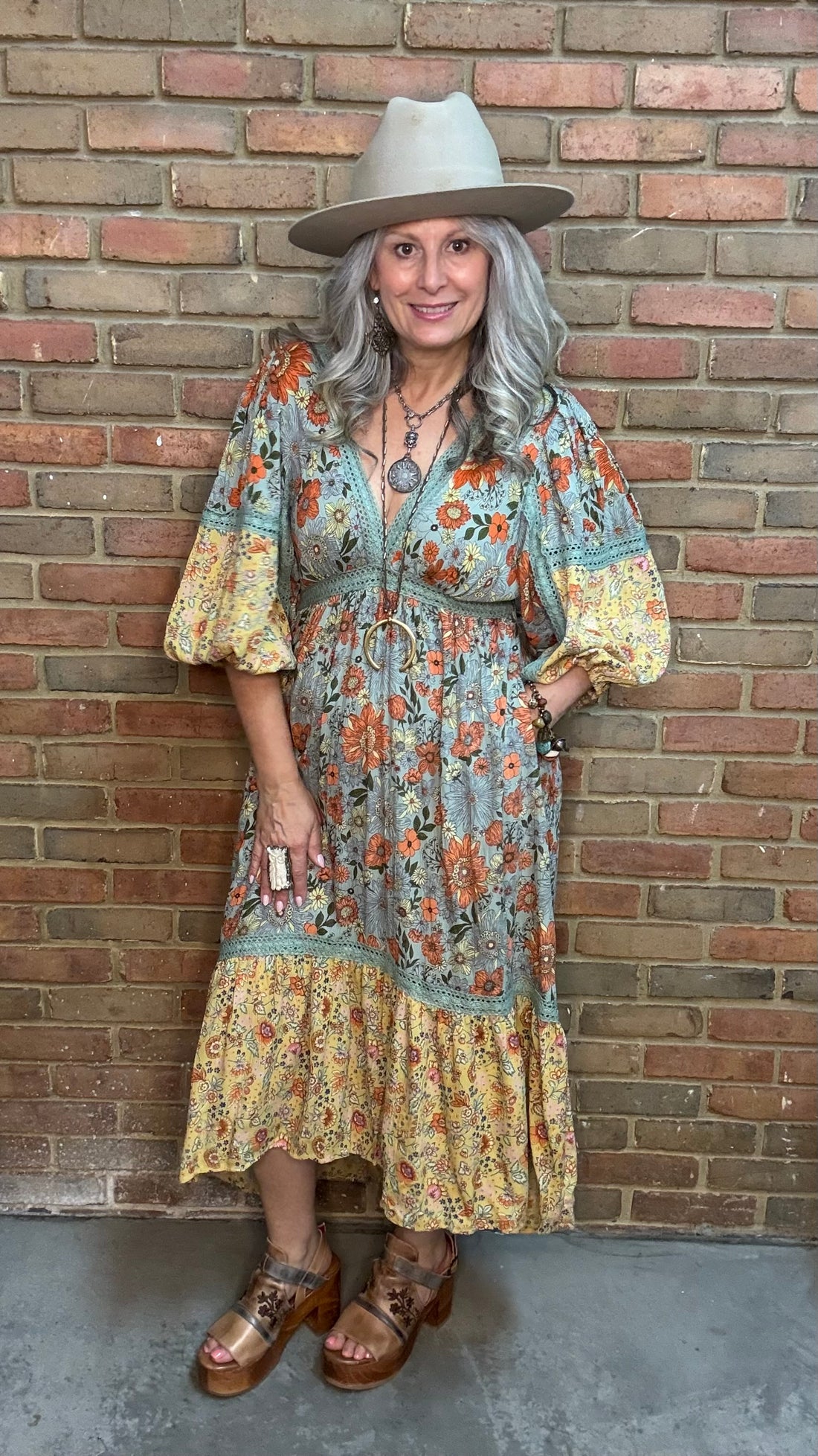 A woman stands against a brick wall wearing a bohemian-style midi dress, the  Long Sleeve V-Neck Hi-Lo Dress, with a blue-gray base and orange floral print, a wide-brimmed beige hat, and brown platform sandals.