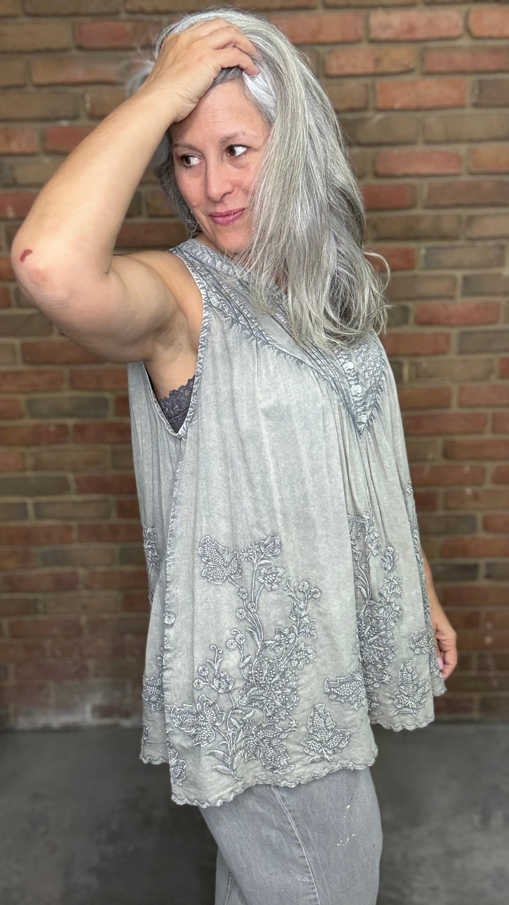 Woman wearing the Sage Floral Embroidery Tank Top, a light-colored sleeveless top with a pattern against a brick wall.