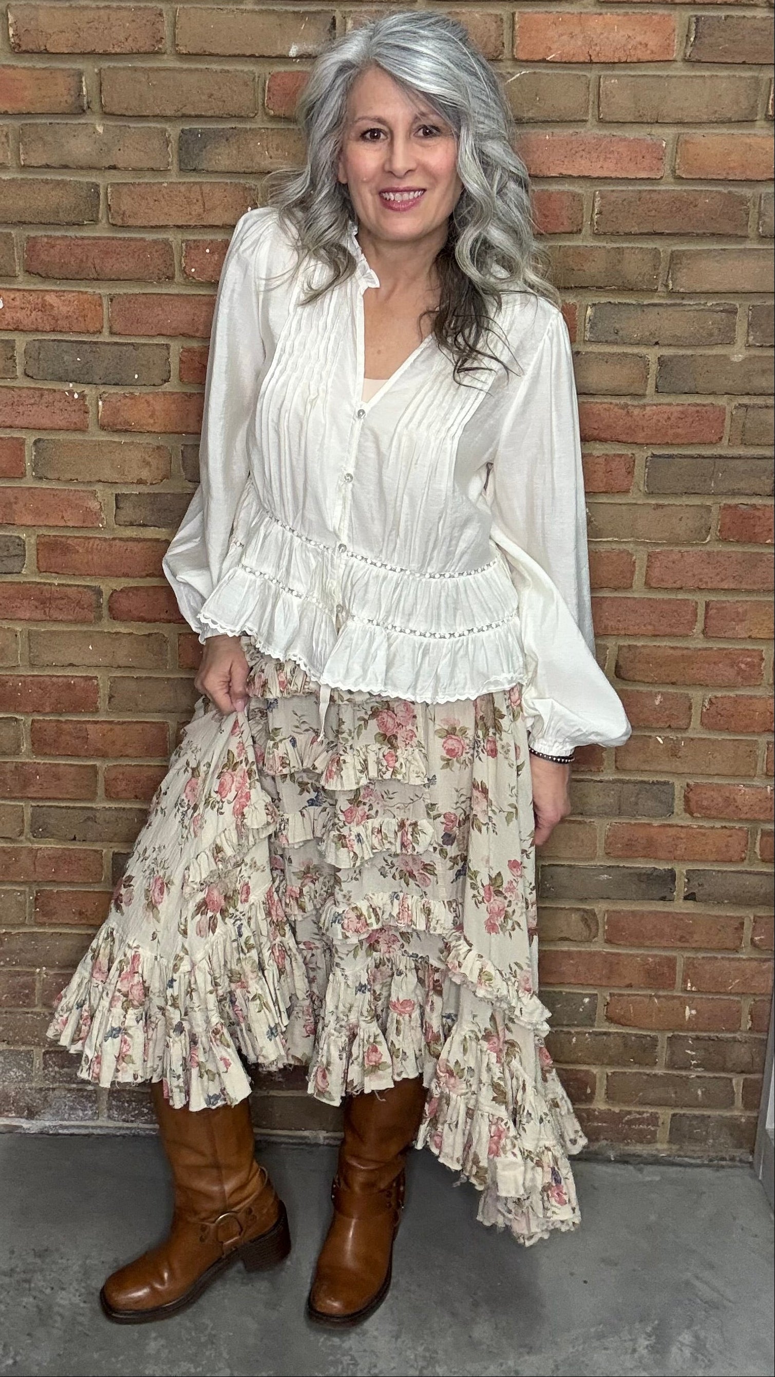 Woman in a white blouse, blooming romance skirt, and Kate Harness Frye boots standing against a brick wall.