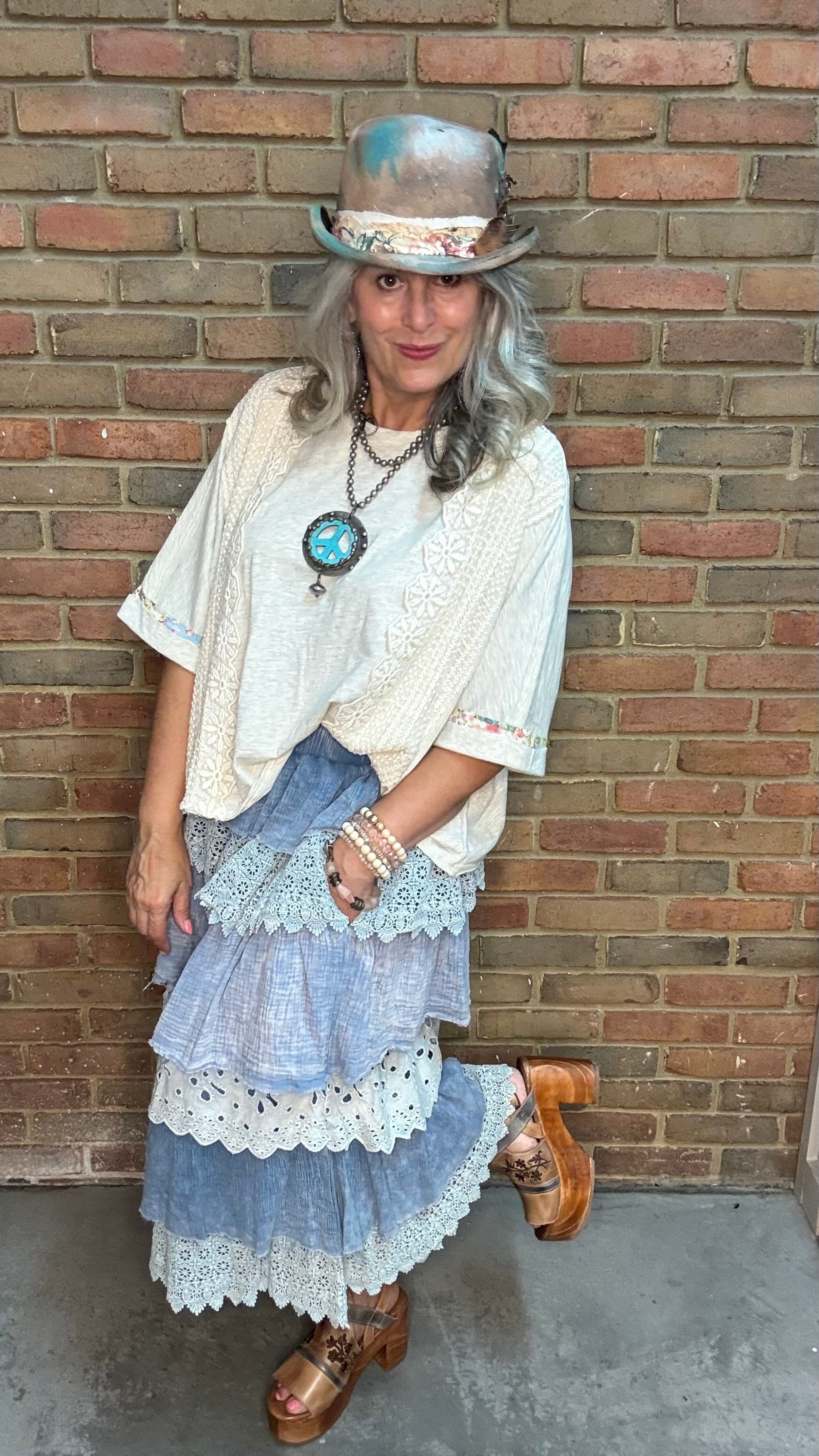 A woman wearing a bohemian-style outfit consisting of a cream lace blouse, a tiered light blue pant with white lace trim, a blue hat, and brown leather shoes, standing against a brick wall.