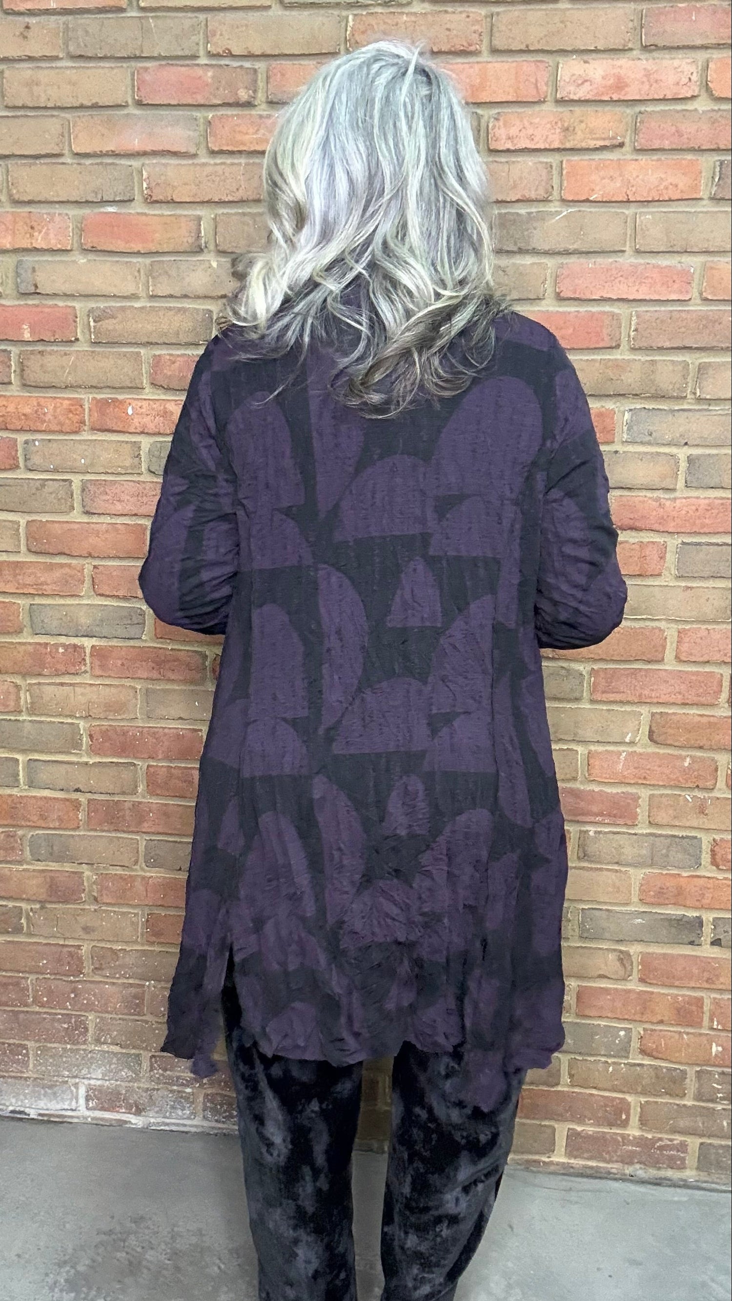 Person wearing a dark patterned outfit standing against a brick wall.