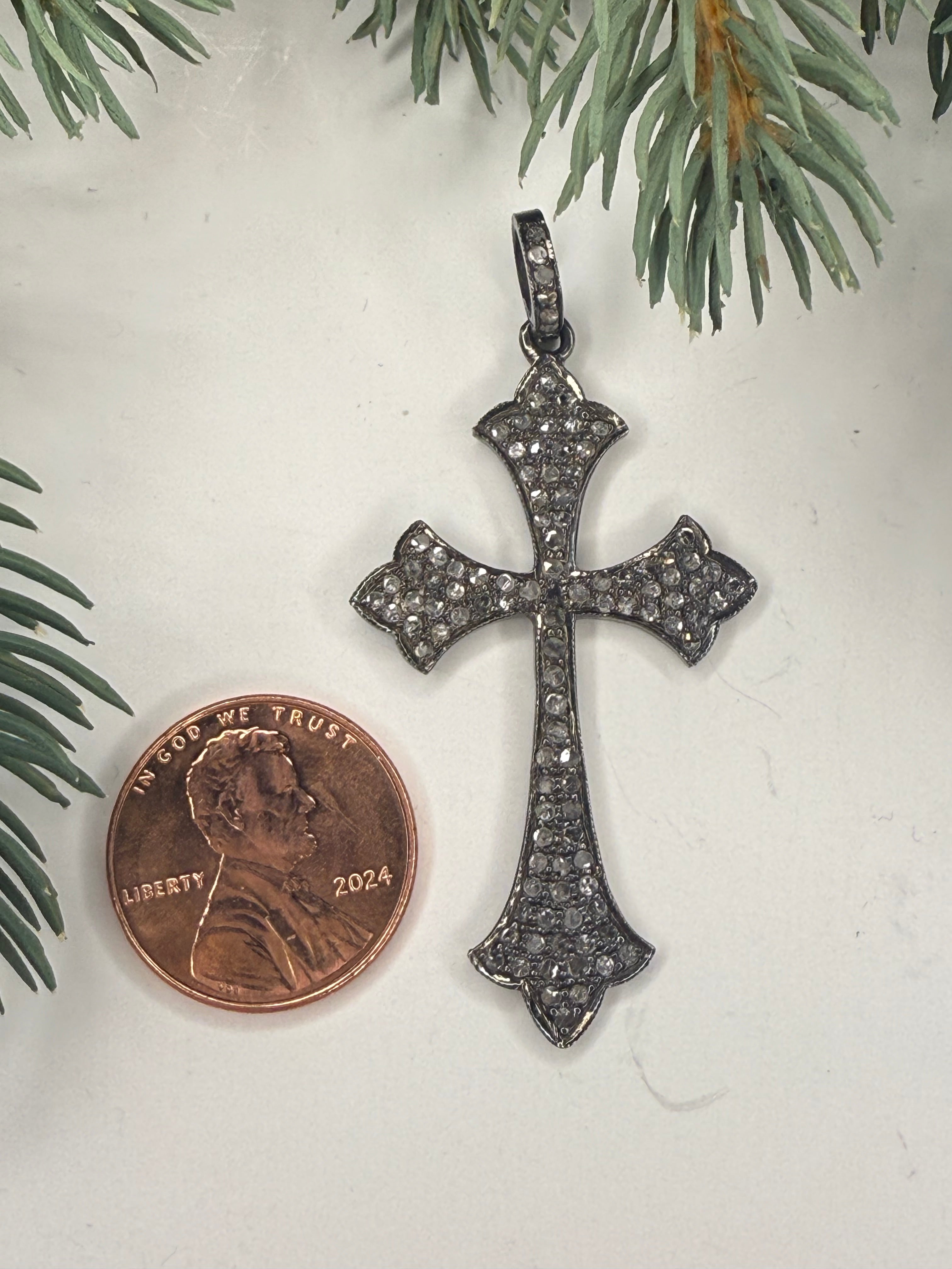Cross shaped pendant next to a penny on a light background with greenery