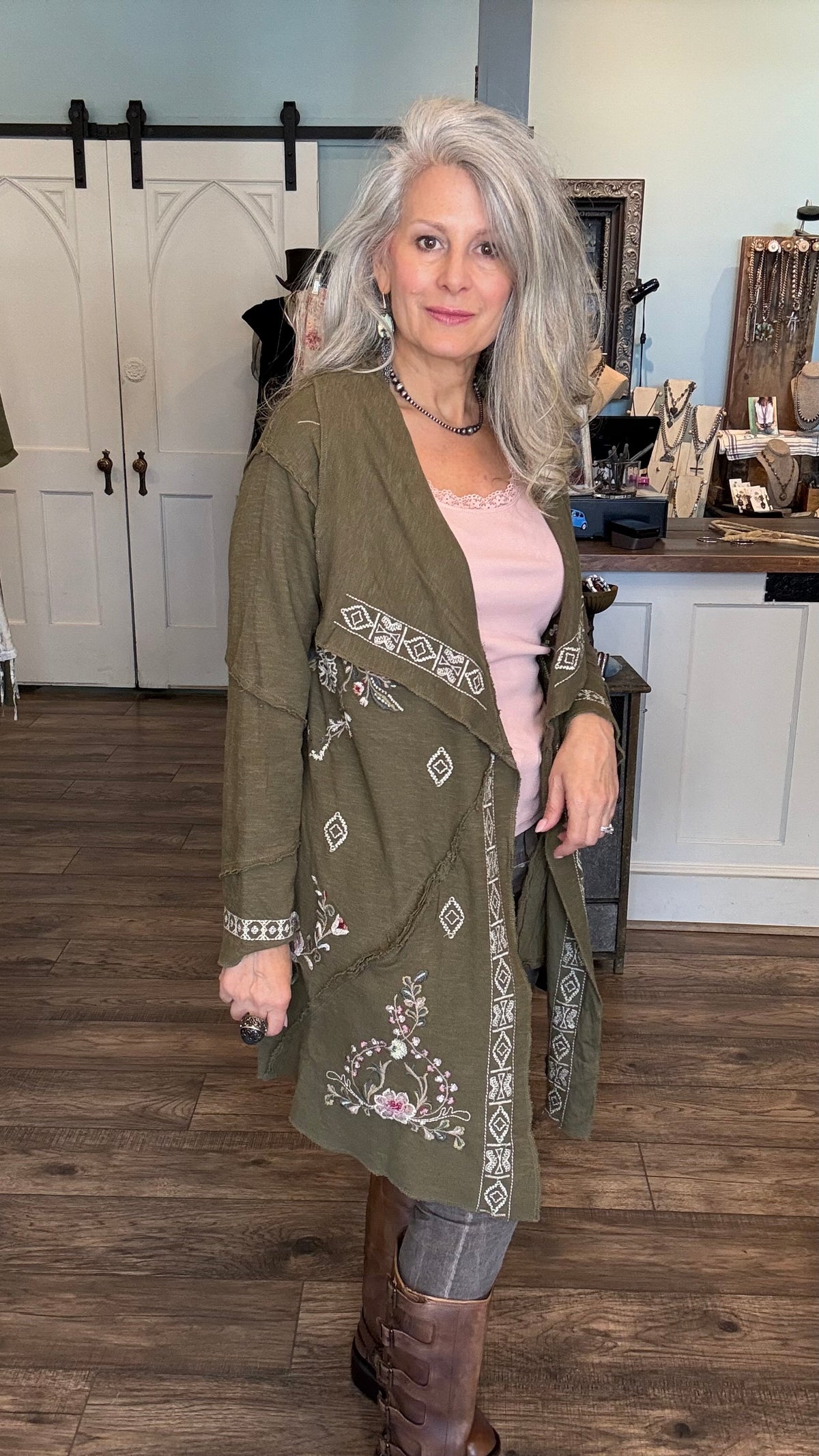 A woman with gray hair wearing an olive green embroidered cardigan over a light pink top and brown boots, standing in a rustic indoor setting with wood floors and white barn-style doors in the background.