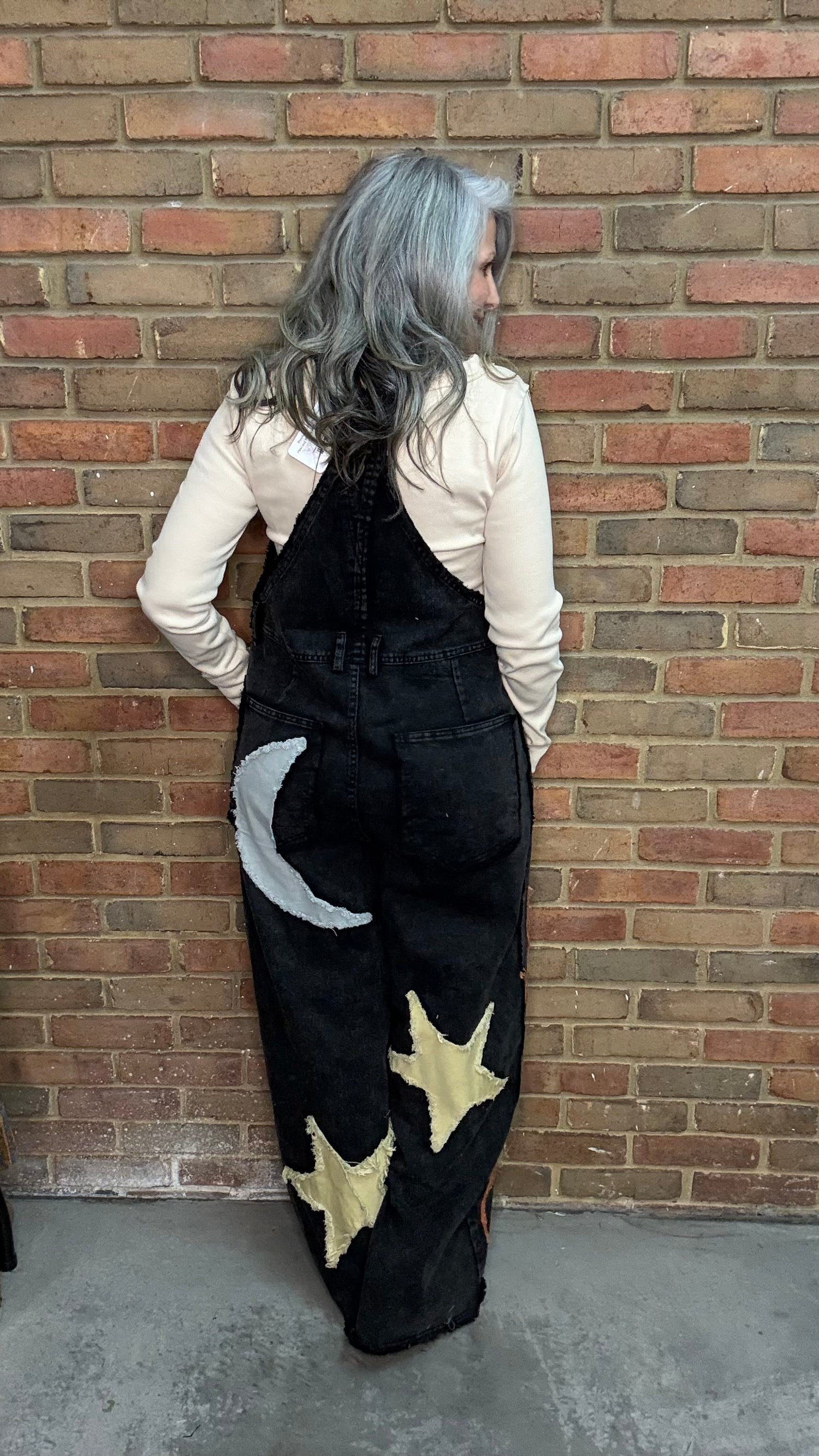 A person with long silver-gray hair stands with their back to the camera, wearing black denim overalls decorated with a white crescent moon and yellow stars, over a cream long-sleeve shirt, set against a red brick wall.