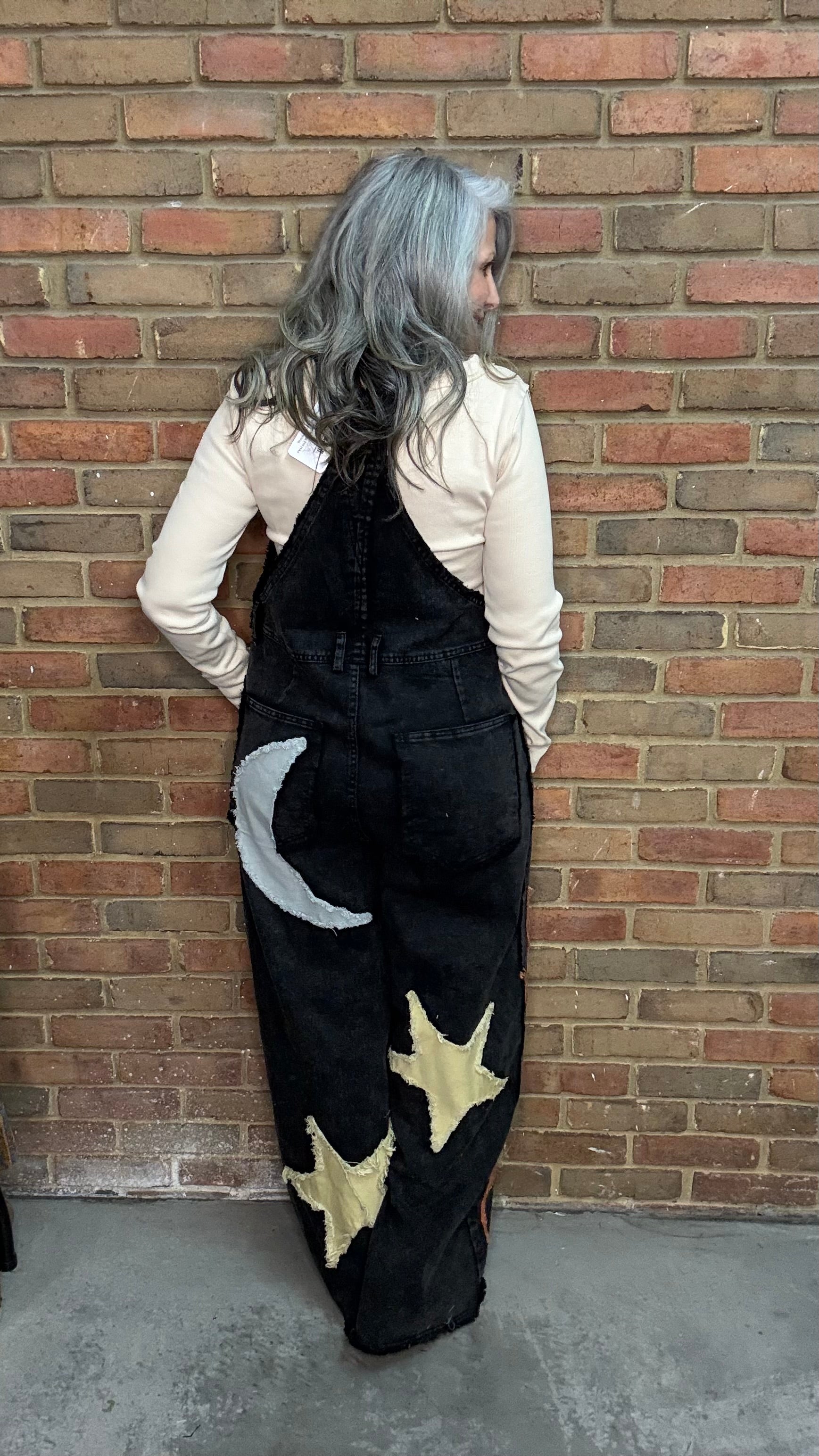 A person with long silver-gray hair stands with their back to the camera, wearing black denim overalls decorated with a white crescent moon and yellow stars, over a cream long-sleeve shirt, set against a red brick wall.