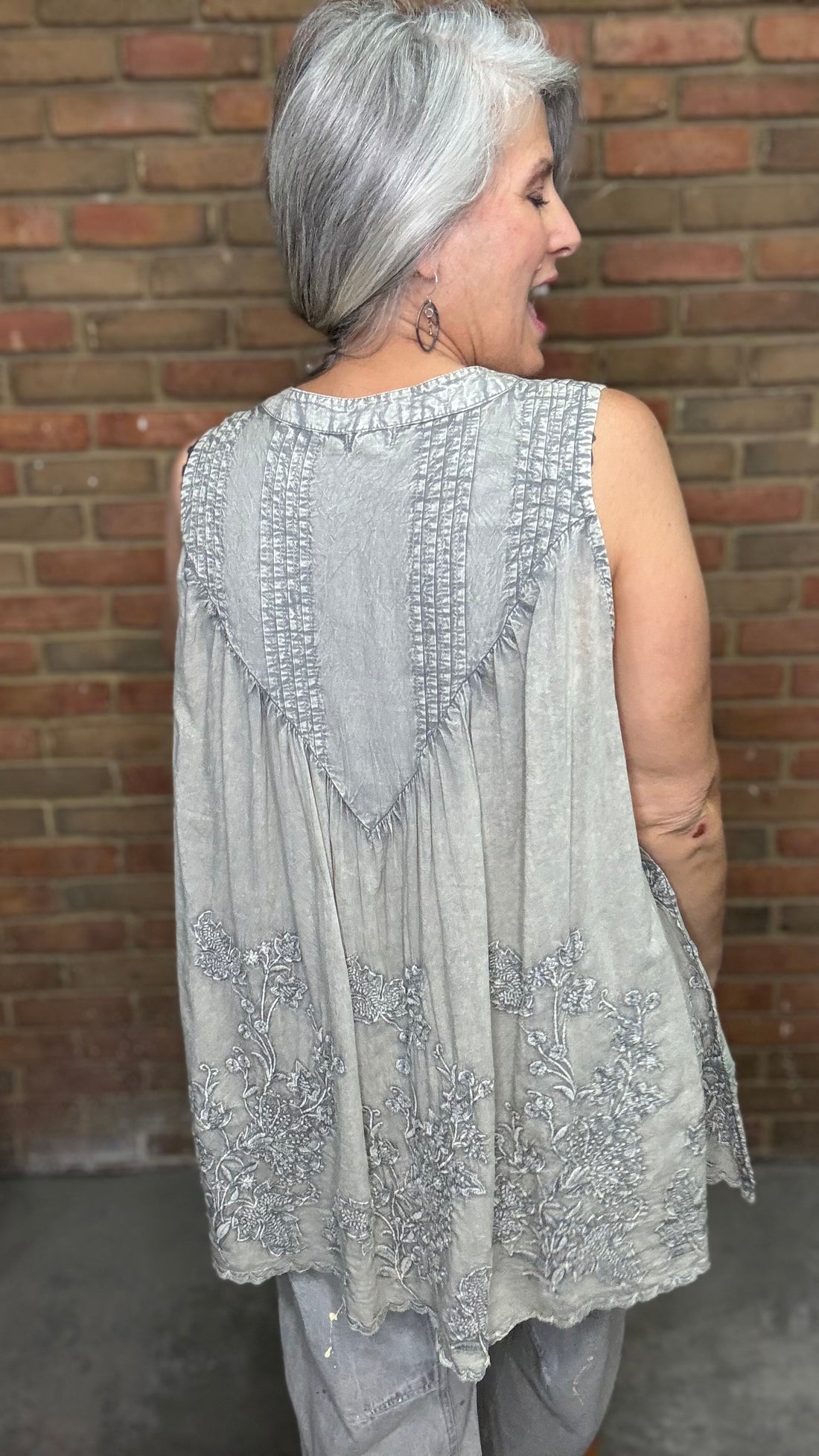 Woman wearing the Sage Floral Embroidery Tank Top, a light-colored sleeveless top with a pattern against a brick wall.