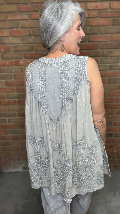 Woman wearing the Sage Floral Embroidery Tank Top, a light-colored sleeveless top with a pattern against a brick wall.