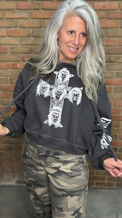 A woman with long gray hair wearing a dark gray graphic sweatshirt with a large white cross and skull print, paired with camouflage pants, standing in front of a brick wall.