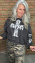 A woman with long gray hair wearing a dark gray graphic sweatshirt with a large white cross and skull print, paired with camouflage pants, standing in front of a brick wall.