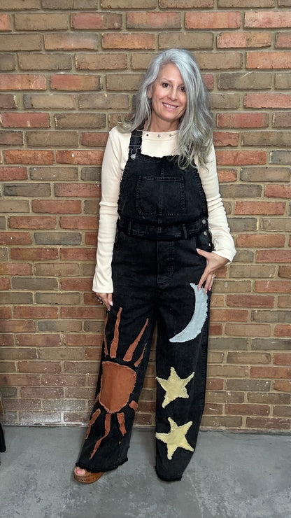 A woman with gray hair stands against a brick wall wearing black denim overalls with embroidered celestial and sun motifs, layered over a cream long-sleeve top.