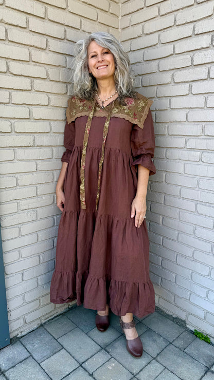 Woman in a brown dress with floral details standing against a white brick wall.