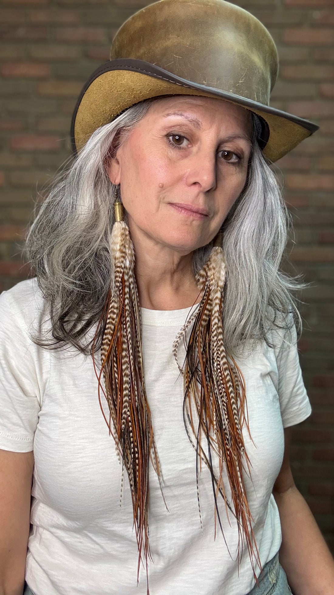 Woman with long feather earrings wearing a leather top hat against a brick wall.
