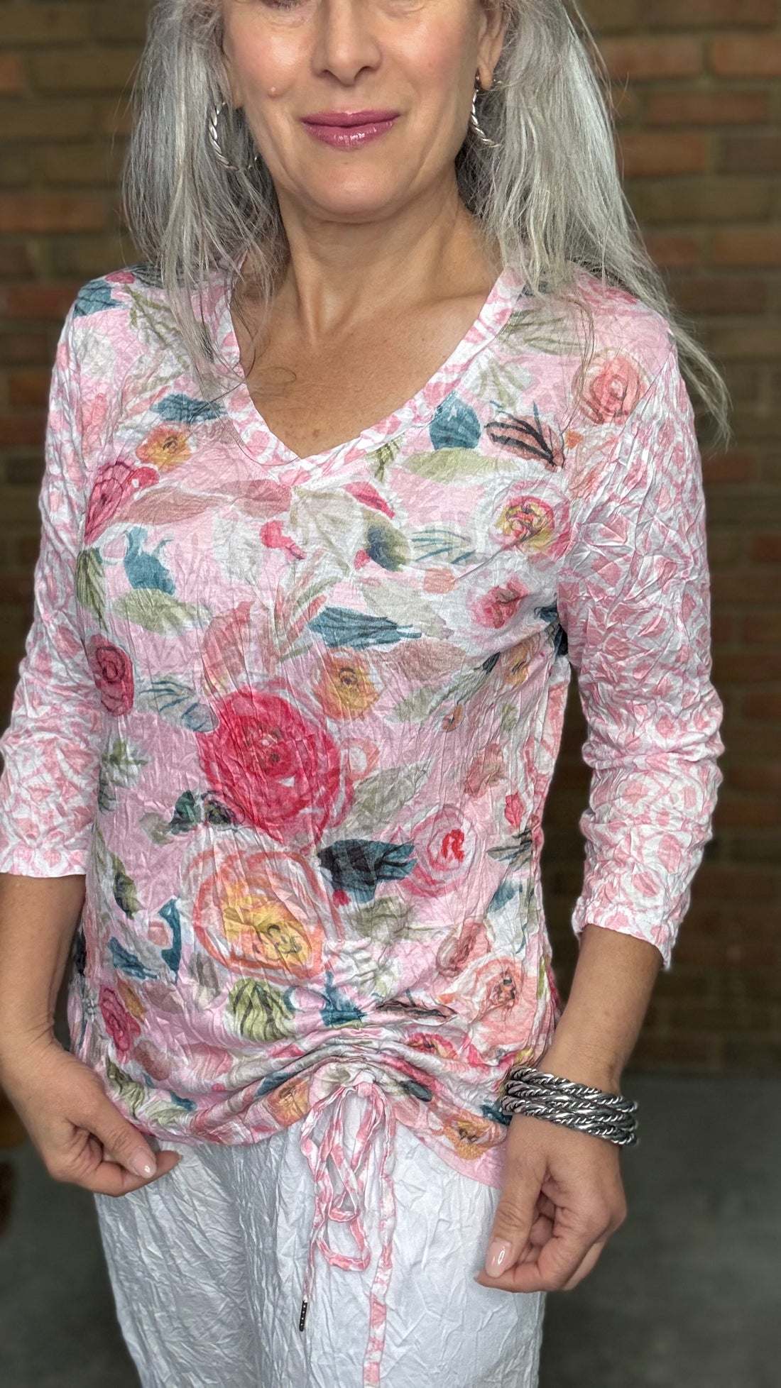 Woman standing against a brick wall wearing the Pink Floral Cinch Side Top and white pants.
