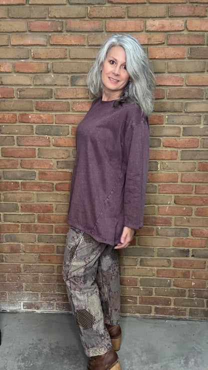 Woman wearing a purple shirt and patterned pants leaning against a brick wall.