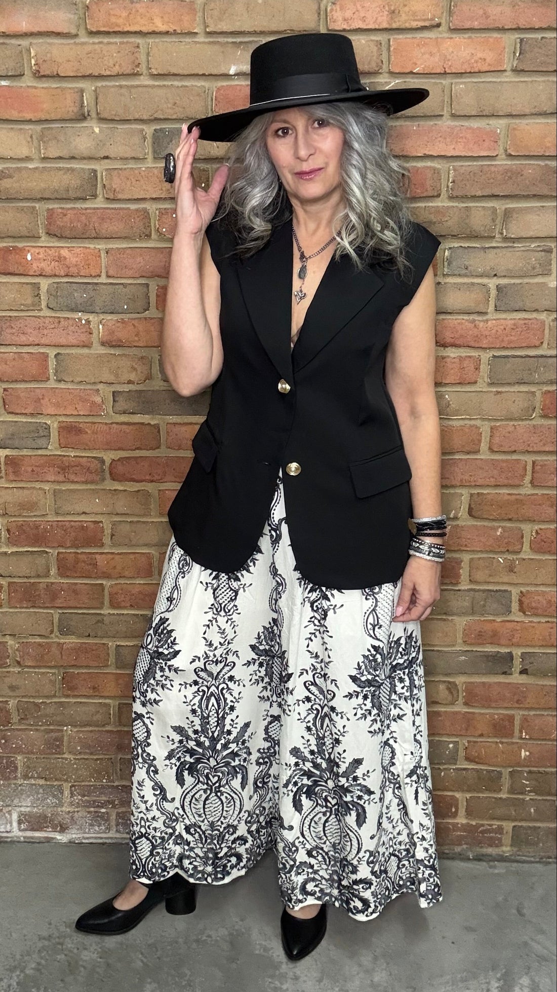 Person wearing a black vest, Floral Print Wide Leg Pant, and a black hat against a brick wall.
