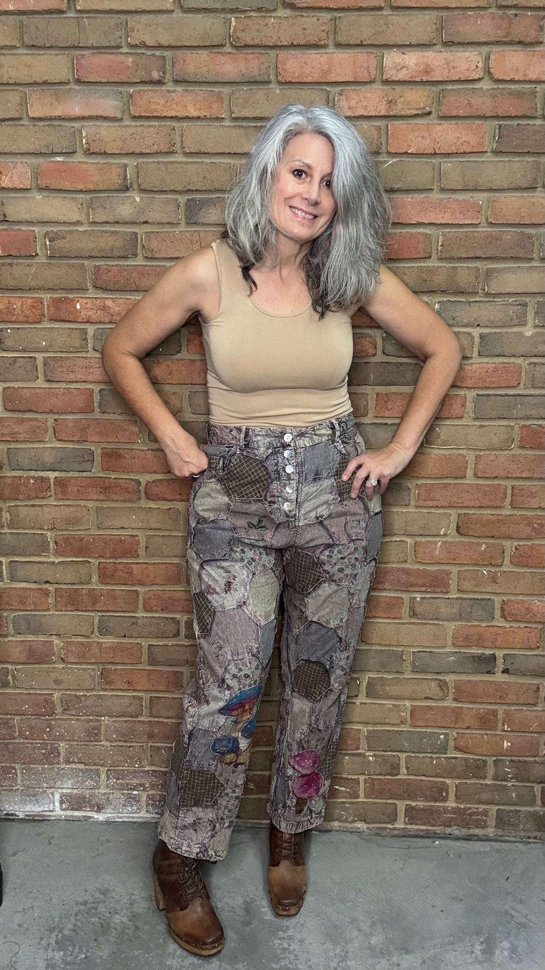 Person wearing a beige tank top and patterned pants leaning against a brick wall.