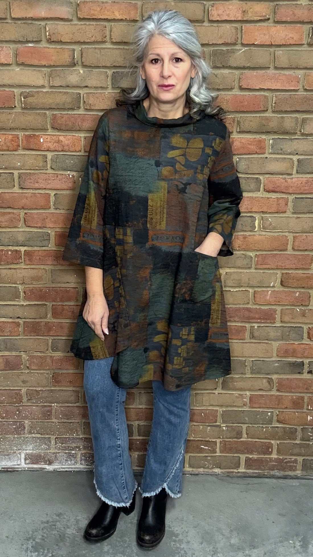 Woman wearing Blythe Tawn Tunic and  the Front Tulip Hem Bootcut Pant jeans standing against a brick wall.