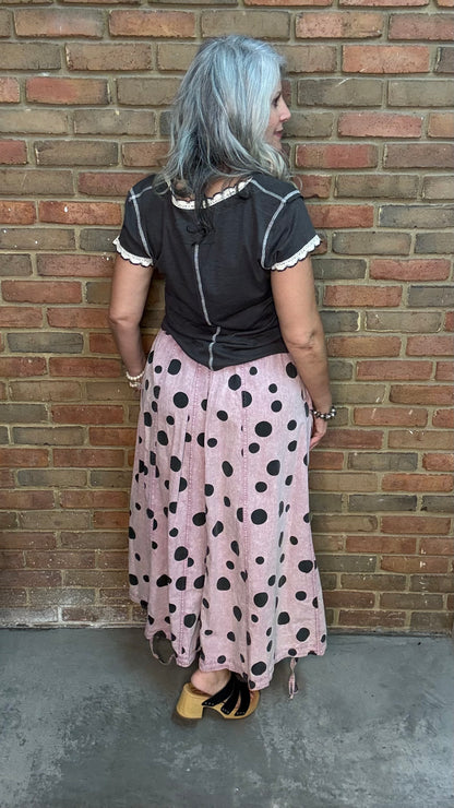 A woman with silver hair stands with her back to the camera, wearing a black short-sleeve top with white trim and a pink maxi skirt with black polka dots, posing in front of a brick wall.
