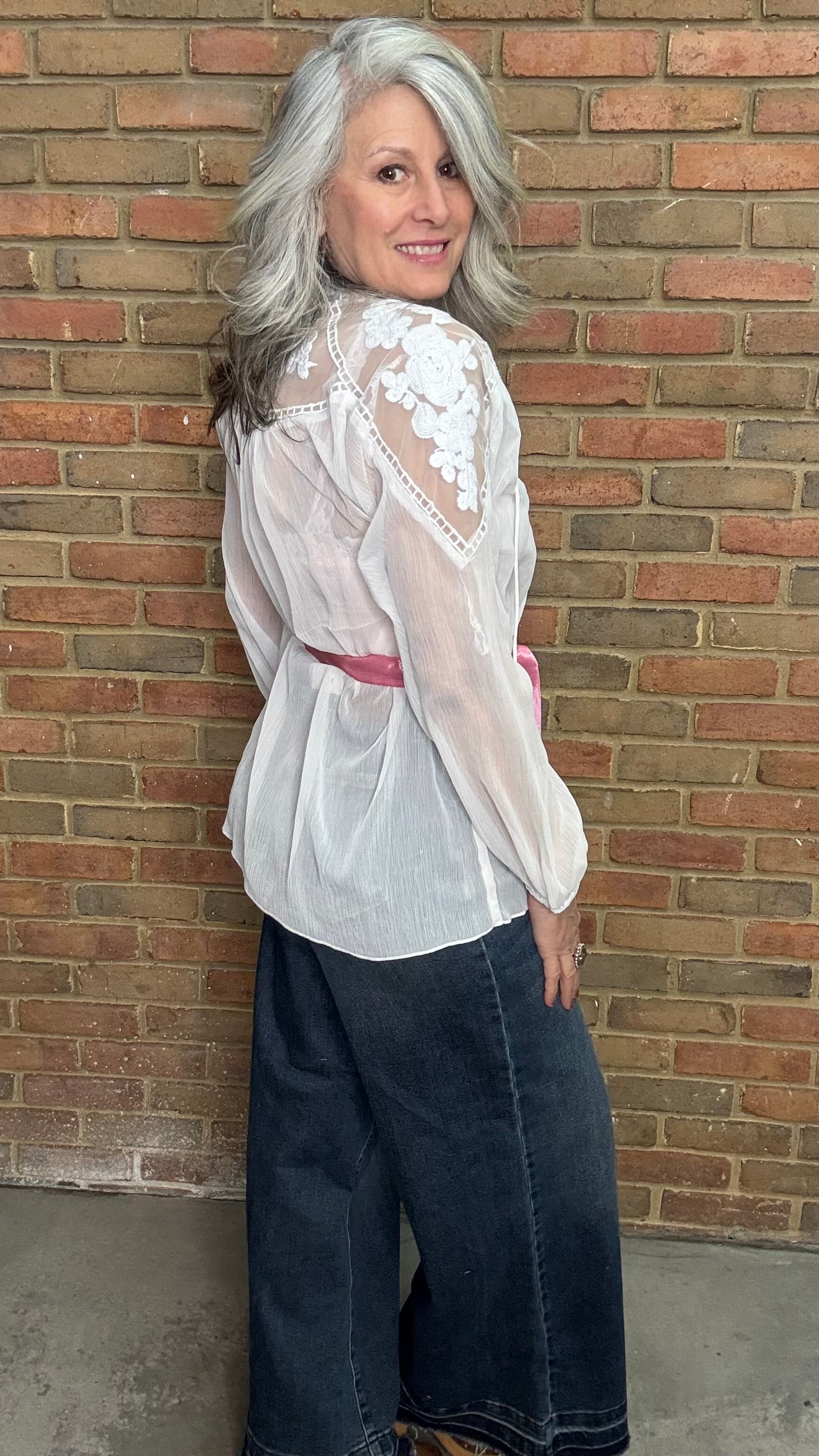 Woman wearing a white blouse with lace details and dark jeans standing against a brick wall.