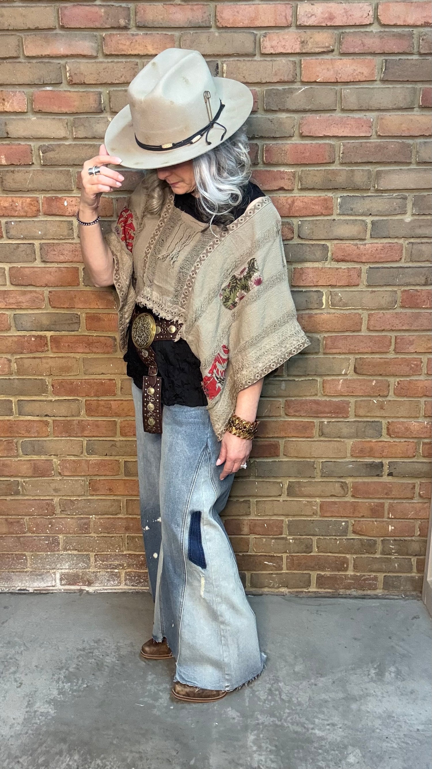 Woman wearing Garden Drift Top with a hat, jeans, and belt against a brick wall.