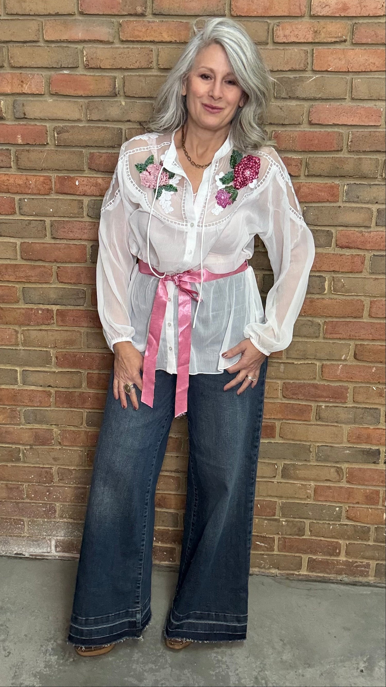 Woman wearing a white blouse with floral embroidery and blue jeans against a brick wall.