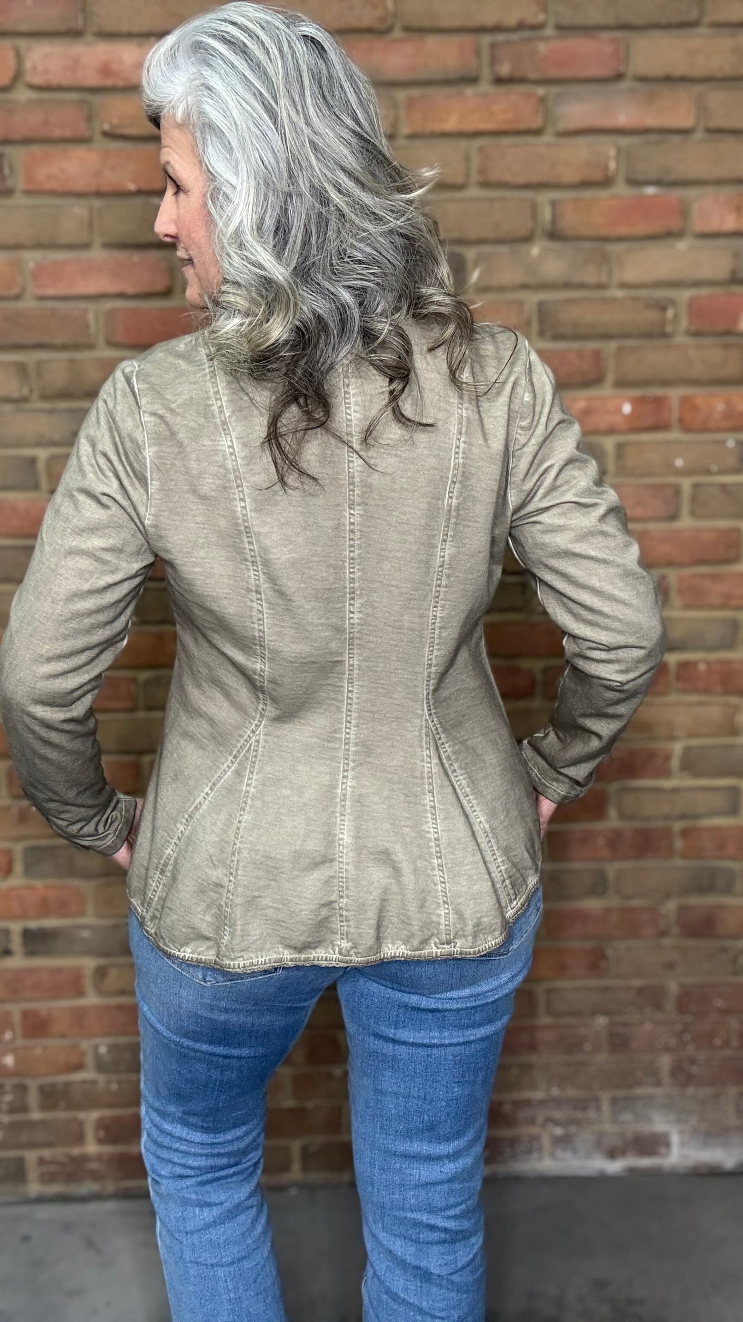 Woman wearing the Colonel Jacket in Taupe with a Sacred Heart Charm Necklace standing in front of brick wall.