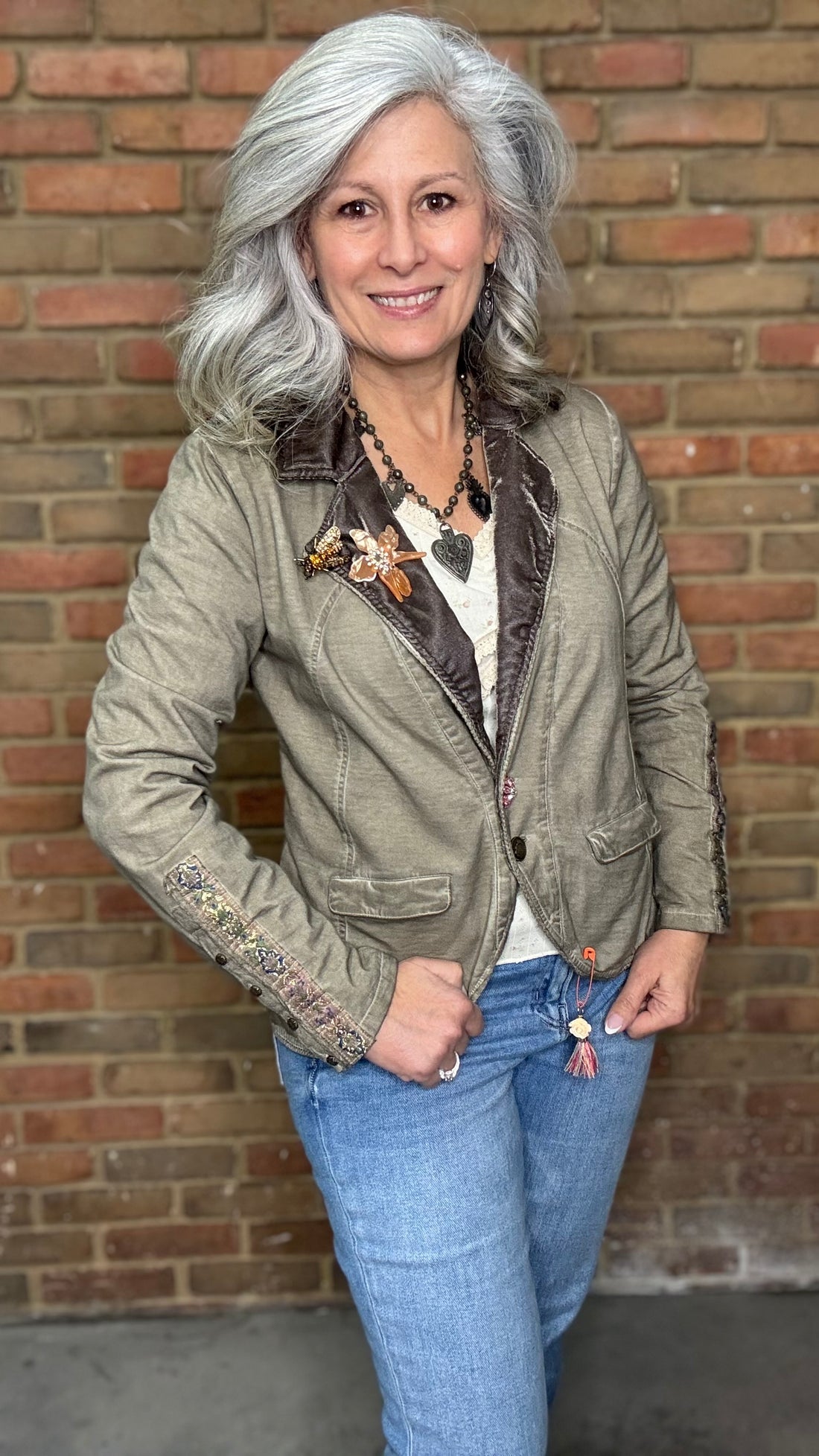 Woman wearing the Colonel Jacket in Taupe with a Sacred Heart Charm Necklace standing in front of brick wall.